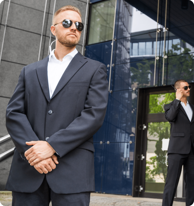 Two men stand guard outside a building wearing dark suits, mirror lens sun glasses, and an ear piece. One is using a walkie talkie