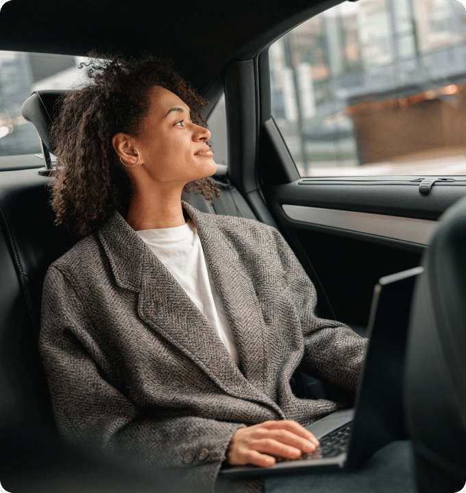 Woman in a backseat of a car with a laptop in her lap, smiling and looking out of the window